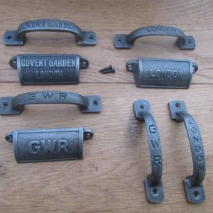 May include: A collection of dark grey cast iron hardware including four arched drawer pulls and three cup pulls. The pieces feature embossed text reading Covent Garden, London, or GWR. Two small black screws are included with the metal hardware.