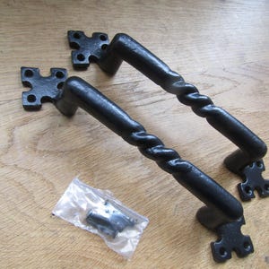 May include: Two black metal door handles featuring a twisted centre design and square mounting plates with four screw holes each. A small clear plastic bag containing matching black mounting screws is positioned between the two handles.
