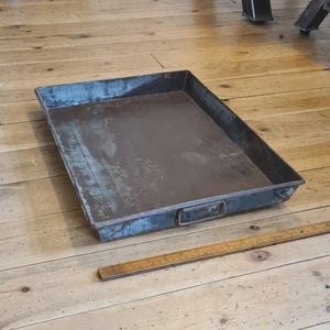 May include: A rectangular, dark-toned metal tray with a weathered, distressed finish and low sides. The tray features a small handle on the side and is positioned next to a wooden ruler that shows the tray is approximately 46 centimetres in length.
