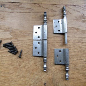 Op de afbeelding: Een set van drie zilverkleurige metalen scharnieren met decoratieve knoppen en een verzameling kleine zwarte schroeven. De scharnieren zijn voorzien van meerdere voorgeboorde gaten voor montage en hebben een verweerde, metalen afwerking.