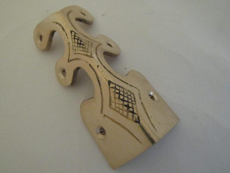 May include: A gold-toned metal corner bracket featuring an elongated, decorative shape with curved edges and multiple circular screw holes. The surface is accented with engraved, dark-filled cross-hatch patterns within diamond-shaped outlines.