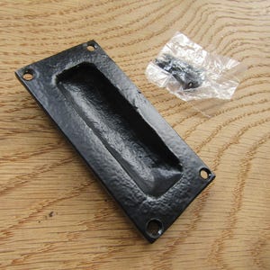 Puede incluir: Un tirador empotrado rectangular de color negro con acabado texturizado y cuatro orificios de montaje en las esquinas. El tirador presenta una zona de agarre central rebajada y se acompaña de una pequeña bolsa de plástico con dos tornillos.