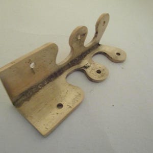 May include: A brass-colored metal hinge with a distinct L-shaped profile and a decorative, scalloped edge featuring three rounded protrusions. The hardware includes four pre-drilled circular mounting holes for installation on a flat surface.