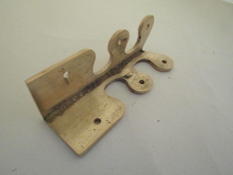May include: A brass-colored metal hinge with a distinct L-shaped profile and a decorative, scalloped edge featuring three rounded protrusions. The hardware includes four pre-drilled circular mounting holes for installation on a flat surface.