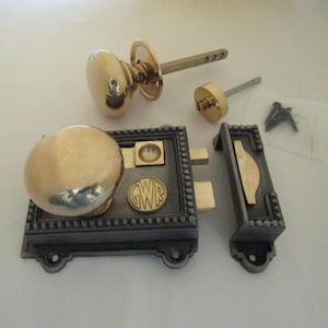 May include: A brass and dark metal rim lock set featuring a round brass knob, a rectangular lock body with a beaded border, a matching strike plate, and a circular emblem embossed with the letters GWR. Includes a separate knob and screws.