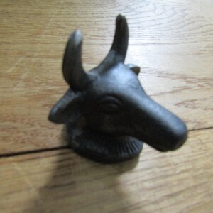 May include: A dark grey, cast metal figurine shaped like a bull head with two prominent, upward-curving horns, pointed ears, and a textured base. The object features a matte finish and a detailed, sculpted snout and eye area, presented in a profile view.
