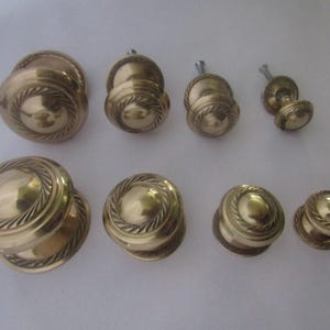 May include: A collection of eight round brass cabinet knobs arranged in two rows, each featuring a smooth domed center surrounded by a decorative twisted rope border. The knobs vary in size, with the largest positioned on the left and the smallest on the right, all showing a polished metallic finish.