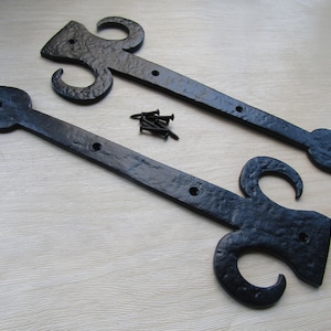 Puede incluir: Dos bisagras de correa de metal negro con un acabado texturizado y un diseño de flor de lis en un extremo, con orificios pretaladrados para el montaje. Un pequeño montón de tornillos negros a juego se encuentra entre las dos bisagras.
