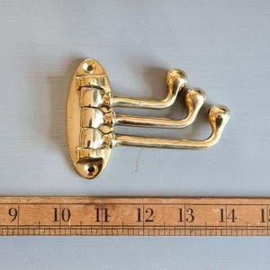 May include: A polished brass triple-arm wall hook with a vertical oval mounting plate featuring two screw holes. The three hinged arms extend horizontally, each ending in a rounded knob. A wooden ruler is positioned below for scale.