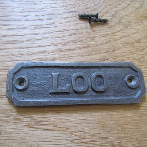 May include: A rectangular cast iron sign with a dark gray, textured finish featuring the word LOO in raised, embossed lettering. The sign includes two pre-drilled holes for mounting and is accompanied by two small, dark metal screws for installation.
