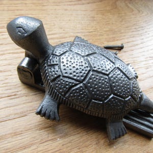 May include: A dark gray cast iron door knocker shaped like a turtle with a textured shell and a hinged head mechanism. The turtle features a detailed, bumpy carapace and four flippers, accompanied by a single metal screw for installation.