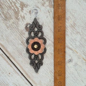 May include: A decorative metal drawer pull with an elongated, ornate dark gray frame and a central peach-colored flower-shaped accent featuring a small gold-toned center. A wooden ruler is placed alongside, showing a length of approximately 4.5 inches.