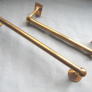 May include: Two polished brass towel rails featuring cylindrical rods with rounded ends. Each rail is supported by two mounting brackets, one with a circular base and the other with a rectangular plate, both finished in a matching metallic gold tone.