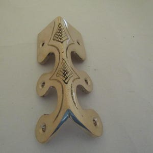 May include: A polished brass corner protector featuring an elongated, decorative shape with six pre-drilled screw holes. The metal surface displays two engraved, triangular grid patterns along its central spine, providing a metallic finish.