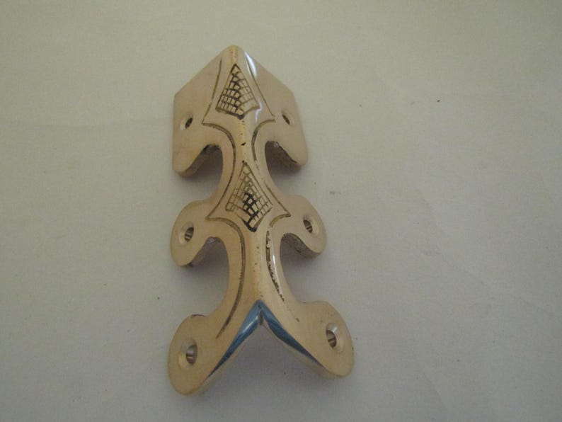 May include: A polished brass corner protector featuring an elongated, decorative shape with six pre-drilled screw holes. The metal surface displays two engraved, triangular grid patterns along its central spine, providing a metallic finish.