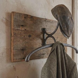 Puede incluir: Un perchero de pared con una pieza rectangular de madera marrón desgastada y una estructura de metal oscuro. Una gorra plana de cuero marrón descansa en el gancho superior, mientras que un blazer de tweed verde oliva cuelga de la barra.