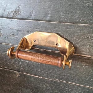May include: A wall-mounted toilet roll holder featuring a polished brass metal bracket with two visible screw holes and a cylindrical wooden roller. The brass hardware has a reflective finish, while the wooden component has a natural brown grain.