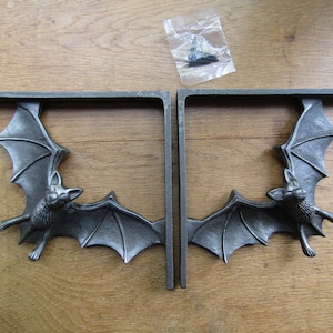 May include: A pair of L-shaped shelf brackets made of dark grey metal, each featuring a sculpted bat with outstretched wings integrated into the frame. A small clear plastic bag containing metal mounting screws is included with the brackets.