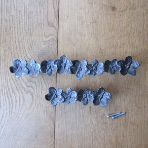 May include: Two dark gray metal drawer pulls featuring a repeating floral design. The larger pull consists of seven connected flower shapes, while the smaller pull features four. Two small silver-colored metal mounting screws are included.