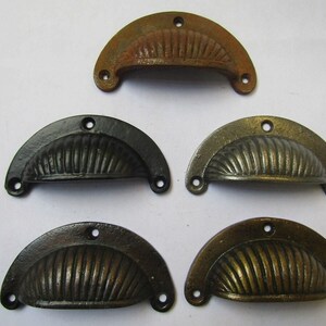 May include: Five semi-circular metal drawer pulls arranged in a group. Each pull features a ribbed, shell-like design with three mounting holes. The finishes vary in colour, including a rusted brown, a matte black, and three metallic bronze tones.