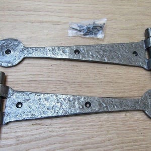 Op de afbeelding: Twee donkergrijze metalen T-hengen met een gehamerde, getextureerde afwerking en een spade-vormig uiteinde. Elke heng heeft een rechthoekige montageplaat met vier schroefgaten en een lange arm met twee extra schroefgaten voor bevestiging.