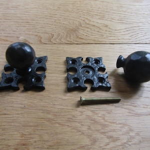 May include: Two black metal spherical knobs with matching square decorative backplates featuring four screw holes each. One knob is attached to a backplate, while the other is detached. A single metal screw is positioned between the components.