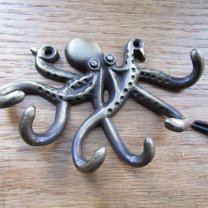 Op de afbeelding: Een donkerbronskleurige metalen wandhaak in de vorm van een octopus, waarbij de gebogen tentakels dienen als individuele haken. De octopus heeft een textuur met kleine cirkelvormige inkepingen langs de armen en twee zwarte schroeven.