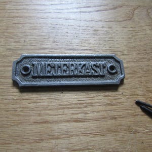 May include: A rectangular cast metal plaque with a textured background and raised lettering that reads METERKAST. The plaque features two circular mounting holes on the left and right sides, and it is accompanied by two small black metal screws.