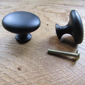 Könnte beinhalten: Zwei mattschwarze Schrankknöpfe mit einer glatten, abgerundeten pilzförmigen Oberseite und einem konischen Sockel. Ein Knopf steht aufrecht, während der andere auf der Seite liegt. Eine silberfarbene Metallschraube zur Montage ist enthalten.