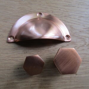 Puede incluir: Un tirador de concha de cobre pulido con tres orificios para tornillos y dos pomos de armario hexagonales con un acabado de cobre a juego. El juego incluye un tirador semicircular y dos pomos de diferentes tamaños, todos con superficie metálica.