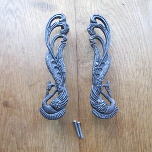 May include: Two dark gray metal door handles featuring a detailed relief design of a peacock with long, flowing tail feathers that curve upward. The handles have a textured finish and are accompanied by two small metal screws for installation.