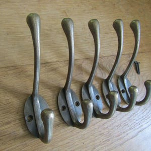 Op de afbeelding: Een set van vijf wandhaken van antiek messingkleurig metaal, elk met een ovale basis met twee schroefgaten, een lange gebogen bovenarm en een kortere onderste haak. Een enkele kleine zwarte metalen schroef ligt bij de meest rechtse haak.