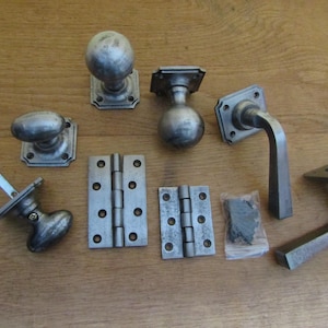 May include: A collection of silver-toned metal hardware including two round door knobs, two lever door handles, two rectangular hinges with screw holes, and a small clear plastic bag containing several dark metal screws for installation.