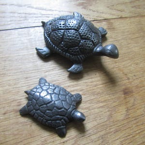 May include: Two dark gray metal turtle figurines with textured shells. The larger turtle features a detailed, perforated shell pattern, while the smaller turtle has a smooth, segmented shell design. Both figures have four legs and a head.