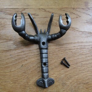 May include: A dark grey, cast iron wall hook shaped like a lobster with two prominent claws and a segmented body. The hook features two circular mounting holes along the centre of the body and is accompanied by two small, matching black metal screws.
