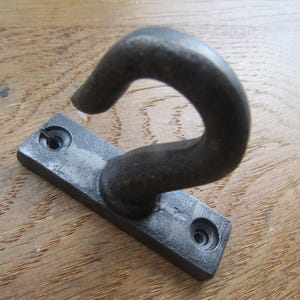 Op de afbeelding: Een donkergrijze metalen wandhaak met een gebogen, J-vormige haak bevestigd aan een rechthoekige montageplaat. De plaat heeft aan beide uiteinden een rond schroefgat en het geheel is afgewerkt met een textuurrijke, matte laag.