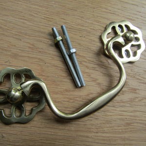 May include: A polished brass drawer pull featuring a curved handle and two decorative circular backplates with openwork floral patterns. The hardware set includes two threaded metal bolts with matching nuts for installation purposes.