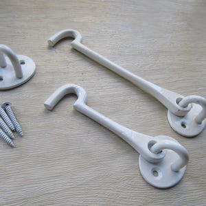 May include: Two white metal cabin hooks with matching mounting plates and a separate eye plate, accompanied by four matching white screws. The hardware features a smooth, glossy finish and a simple, functional design for securing doors or windows.