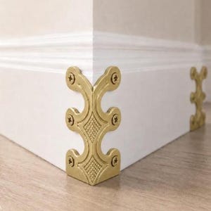 May include: A gold-toned metal corner guard featuring an ornate, curved design with a textured diamond pattern in the center. The piece is secured to the corner of a white baseboard with six visible screws, providing a decorative finish.