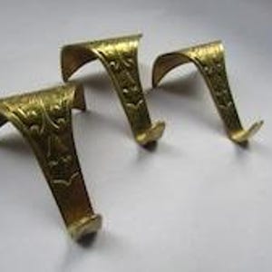 May include: Three gold-colored metal hooks with a curved top design and an embossed decorative pattern along the front surface. Each hook features a pointed end and a wide, flat top section intended for mounting or hanging over a flat edge.