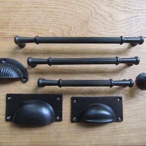 Puede incluir: Una colección de herrajes de metal negro que incluye tres tiradores de barra estriados de diferentes longitudes, un tirador de concha semicircular con crestas verticales, un pomo redondo estriado y dos tiradores de cazoleta rectangulares.