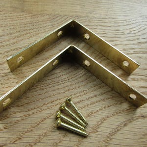 May include: Two L-shaped brass corner braces with pre-drilled screw holes, accompanied by four matching brass screws. The metal brackets feature a smooth, polished finish and are designed for reinforcing right-angle joints in furniture or shelving.