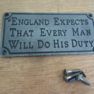 May include: A rectangular grey metal plaque featuring the embossed text "ENGLAND EXPECTS THAT EVERY MAN WILL DO HIS DUTY" with four corner screw holes. Three matching metal screws are positioned below the plaque on the surface.