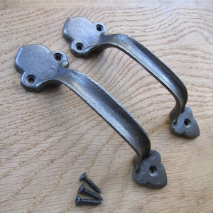 May include: Two dark grey metal door handles with a textured finish and curved design. Each handle features two mounting plates with circular screw holes at the ends. Three small black metal screws are positioned below the handles.