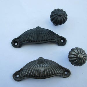 Puede incluir: Dos tiradores de metal oscuro y dos pomos redondos con un diseño acanalado. Los herrajes presentan una forma curva similar a una concha para los tiradores y un patrón circular de pétalos para los pomos, con un acabado metálico oscuro y desgastado.