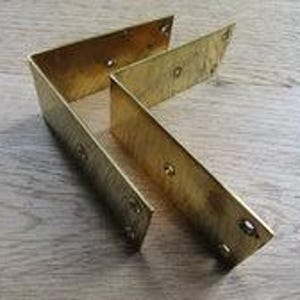 Può includere: Due staffe per mensole a forma di L realizzate in ottone lucido. Ogni staffa presenta un design piatto e angolato con molteplici fori circolari preforati lungo entrambi i lati per il montaggio. La superficie metallica ha una finitura dorata.
