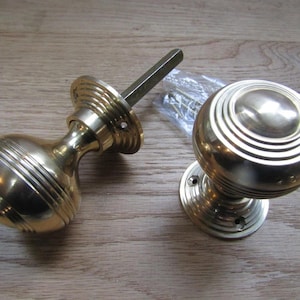 Op de afbeelding: Een paar gepolijste koperen deurknoppen met een bolvorm en concentrische cirkelvormige ribbels. Eén knop is voorzien van een vierkante metalen stift die uit de basis steekt, en een klein plastic zakje met bevestigingsschroeven ligt ernaast.