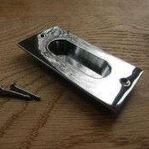 Pode incluir: Um puxador embutido retangular com acabamento em cromo polido e uma pega central rebaixada. O puxador apresenta dois orifícios para parafusos pré-perfurados nas extremidades superior e inferior, acompanhado por dois pequenos parafusos metálicos.