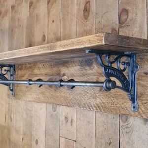 May include: A wall-mounted shelf featuring a natural wood plank surface and a matching backboard. Two ornate black cast iron brackets support the shelf, connected by a horizontal metal rod that functions as a rail for hanging items.