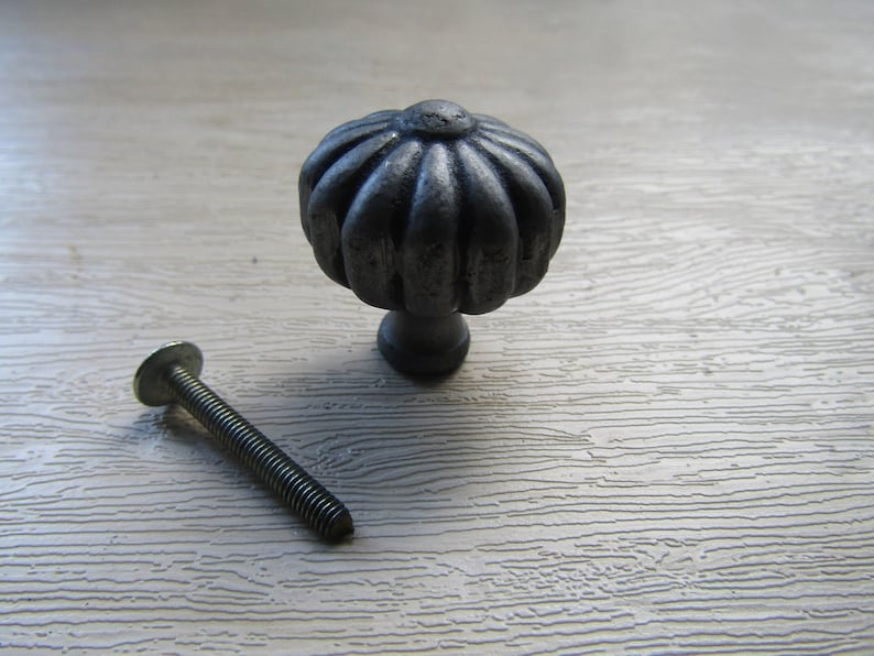 K&ouml;nnte beinhalten: Ein dunkelgrauer M&ouml;belknopf aus Metall mit einer abgerundeten, geriffelten, k&uuml;rbisartigen Form und einer strukturierten Oberfl&auml;che. Der Knopf hat eine mittige, kreisf&ouml;rmige Oberseite und vertikale Rippen. Eine lange Gewindeschraube liegt bei.